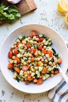 a white bowl filled with cucumber, tomato and chickpea salad next to lemon wedges