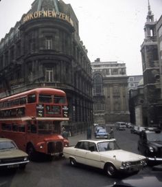 a red double decker bus driving down a street next to tall buildings and parked cars