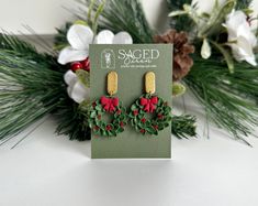 a pair of green and red christmas wreath earrings on top of a card next to flowers