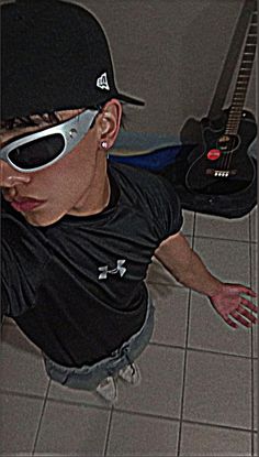 a young man wearing sunglasses and a baseball cap with his hand on the ground next to a guitar