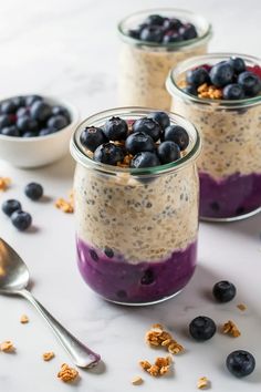 three jars filled with blueberries and granola on top of a white table next to spoons