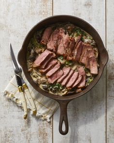 a skillet filled with meat and vegetables on top of a wooden table next to utensils