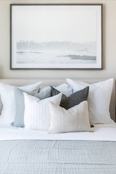 a white bed topped with lots of pillows next to a framed painting on the wall