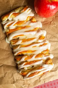 an apple and cinnamon roll with icing next to it on wax paper, one red apple is in the background