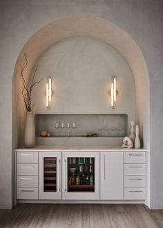 a white cabinet with bottles and glasses in it next to a vase on the counter