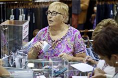 an older woman sitting at a table with other people in the background looking at items on display