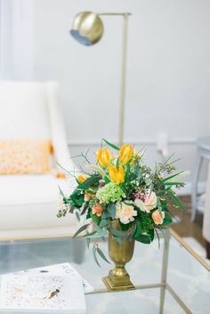 a glass table topped with a vase filled with yellow and pink flowers next to a white chair