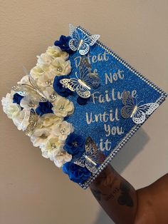 a blue graduation cap with white flowers and butterflies is held up by someone's arm