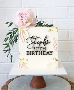 a white cake with pink flowers on top sitting on a wooden stand in front of a brick wall