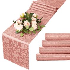 pink table runner with white roses and green leaves on it next to four folded napkins