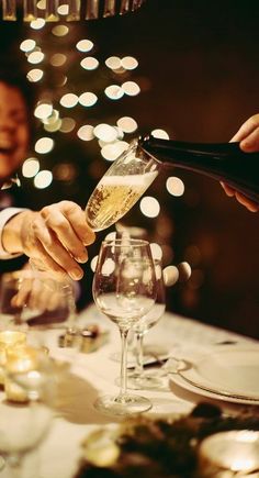 two people pouring champagne into wine glasses at a dinner table with lights in the background