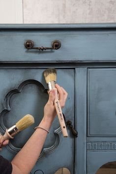 a woman is holding a paintbrush in front of a blue dresser with drawers and knobs