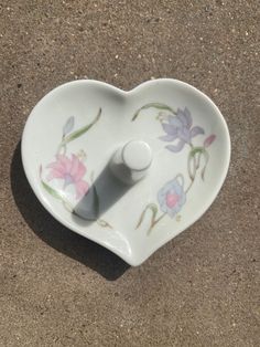 a white heart shaped dish with flowers painted on the front and sides, sitting on a brown surface