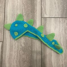 a crocheted blue and green dinosaur hat laying on the floor next to a stuffed animal