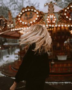 a woman standing in front of a merry go round at the park with her hair blowing in the wind