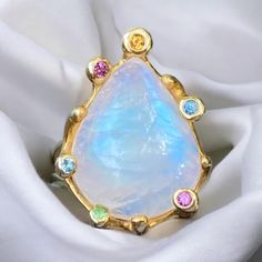 This beautiful  moonstone ring is set in simple 925 sterling silver, 14k yellow gold plated band that splits in two closer to the top with seamless transition into the free form 10 prongs.  Every other prong is decorated with multi color tourmaline dainty crystals elegantly complimenting the majestic moonstone cabochon.  The large crystal's colors show cool blues, gold and even some lilac hues depending on the light, shown in the photos. Very pretty and feminine design. Moon stone deposits are in many countries Armenia, Australia, Mexico, but these stones came from Sri Lanka. Moonstone is the sacred stone of India. Beautiful shades of blue color made this stone so popular, that it gained an alternate name "Rainbow" moon stone. The color of the stone is clear until it is turned under a cert Ring Teardrop, Blue Shades Colors, Moonstone Cabochon, Moonstone Stone, Cabochon Ring, Lilac Color, Large Crystals, Yellow Gold Ring, Moonstone Ring