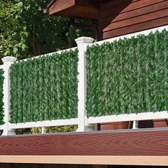 a white fence covered in green plants next to a building with trees and bushes behind it