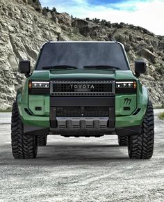 the front end of a green truck parked on top of a dirt field next to a mountain