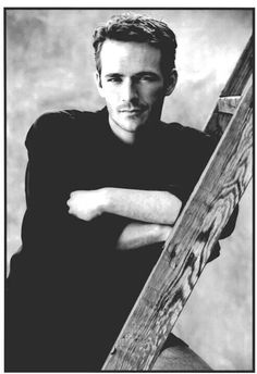 a black and white photo of a man leaning against a wooden fence with his arms crossed