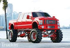 a large red truck parked in front of a body of water with tall buildings in the background