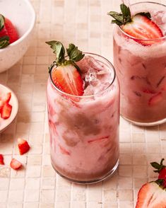 two glasses filled with strawberry milkshakes on top of a tiled counter next to bowls of strawberries