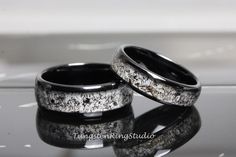 two wedding rings with black and white speckles are sitting on a reflective surface