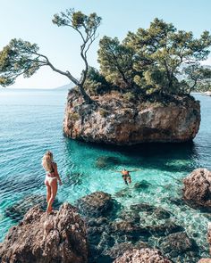 two people are swimming in the water near some rocks and trees, while another person is standing on top of an island