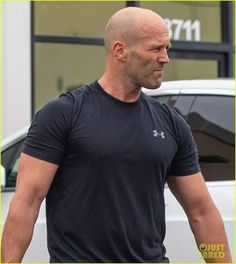 a bald man in a black shirt is walking down the street with his hand on his hip