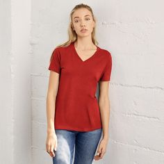 a woman standing in front of a white wall wearing a red shirt and blue jeans