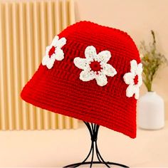 a red crocheted hat with white flowers on it sitting on a stand next to a vase