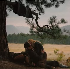 two people sitting on the ground under a tree with mountains in the backgroud