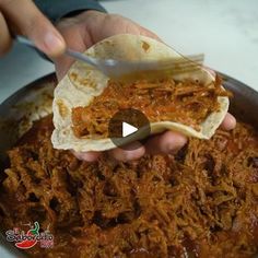 a person holding a tortilla with shredded meat in it and another hand scooping something out