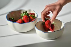 two white bowls with strawberries in them on a table top, one being picked from another bowl