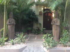 two statues in front of a house surrounded by palm trees and other tropical plants on either side of the door