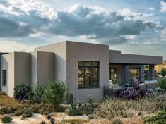 a small house in the middle of a desert with plants and shrubs around it, on a cloudy day