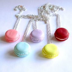 four small macaroons are attached to chains on a white surface, one is pink, one is blue and the other is yellow