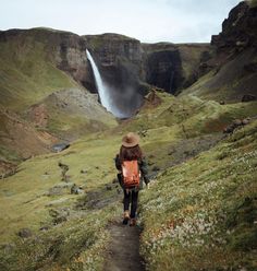 a woman with a backpack walking up a hill towards a waterfall in the distance,