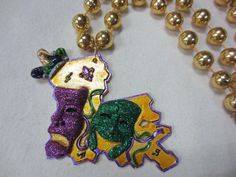 Comedy stands out on this bead as tragedy is cut off the state. This bead features the state of Louisiana in gold with a jester hat and comedy and tragedy in purple and green glitter accents. Medallion is approximately 3" x 3" at the bottom of a 12mm gold beaded 42" long necklace. Available individually, 12-pack, or case pricing (10 dozen/120 pieces) **CLEARANCE PRICING ON 12-PACK PURCHASE** Purple Jewelry For Mardi Gras, Purple Beaded Jewelry For Mardi Gras, Round Beads Jewelry For Mardi Gras Party, Metal Mask, Jester Hat, Feather Mask, New Orleans Mardi Gras, Comedy And Tragedy, Beaded Hat