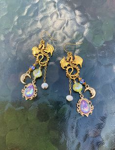 These dragon drop earrings are so much fun! Made with gold tone antique dragons. Lavender and lime green AB crystals along with a crescent moon dangle from its curled tail.  Large, bold and light weight. These are perfect for a fan of fantasy creatures.  They have about a 2.5 inch drop with simple ear wires. Beautiful statement earrings unlike any you have ever seen. Gold Fantasy Jewelry With Dragon Design, Fantasy Gold Jewelry With Dragon Design, Magical Gold Metal Jewelry, Mystical Gold Drop Earrings, Mystical Gold Crystal Earrings Gift, Magical Gold Nickel-free Jewelry, Magical Gold Earrings As Gift, Magical Gold Earrings For Gift, Mystical Handmade Gold Crystal Earrings