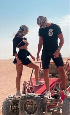 a man and woman standing on top of a red vehicle in the middle of desert