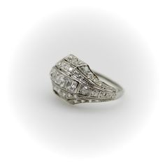 This platinum diamond ring is on the cusp of the Edwardian and Art Deco eras. The centerpiece of the ornate ring are three Peruzzi cut bezel set diamonds —one is an elongated rectangular shape, while the diamonds on either side are truncated, cut specifically for this ring. The larger diamonds are surrounded by a selection of 40 single and Old Mine Cut diamonds that are bead set into the ring. The ring has layers divided by milgrain work that tie together the lace-like patterns of the Edwardian era and the geometric designs of the Art Deco era in alternating bands. There is an added detail of a hand engraved scrolling foliate on each side of the ring. The ring has open work on the bottom that lets in the light and also reveals the triangular bottom shape of the diamonds—a nice touch that h Timeless Square Cut Rose Diamonds Jewelry, Diamond Ring With Baguette Diamonds And Rectangular Stone, Rectangular Diamond White Ring With Baguette Diamonds, Elegant Silver Diamond Ring With Baguette Diamonds, Silver Diamond Ring With Baguette Diamonds, Formal Art Deco Diamond Ring With Baguette Diamonds, Heirloom Diamond Rings With Square Cut, Luxury Diamond Ring With Rectangular Stone For Anniversary, Wedding Square-cut Platinum Diamond Ring