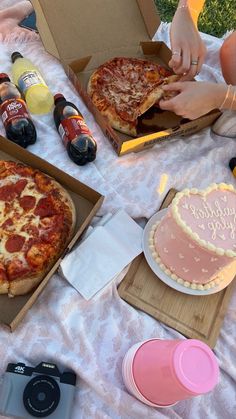 two pizzas in boxes on a table with other food and drink items around them