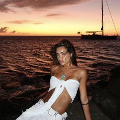 a woman in a white dress sitting on the rocks near the ocean with a sailboat in the background