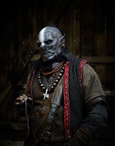 a man dressed up as a demon with beads and necklaces on his neck, standing in front of a wooden wall