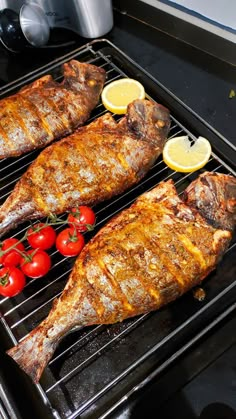 two fish are cooking on the grill with tomatoes and lemons next to each other