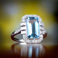 A Geometric ring that features a lively emerald cut natural aquamarine weighing approximately 2.35 carats with a stunning teal saturation. Surrounding this ring is old European cut diamonds and 3 baguette-cut diamonds on each side. This platinum ring has a low profile and has a floral under-gallery design Art Deco Topaz Ring With Center Stone, Art Deco Diamond Emerald Ring Octagon Shape, Art Deco Blue Asscher Cut Ring, Art Deco Emerald Cut Topaz Ring, Art Deco Aquamarine Blue Ring, Art Deco Blue Aquamarine Ring, Blue Aquamarine Art Deco Rings, Art Deco Diamond Topaz Ring, Art Deco Baguette Cut Gemstone Ring