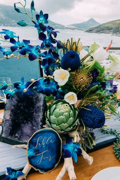 an arrangement of blue flowers on a table near the water with other items in front of it