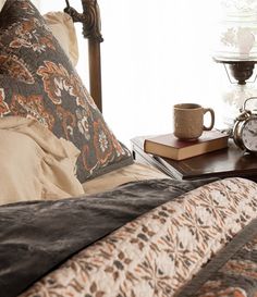 a coffee cup on a table next to a bed with pillows and blankets in front of a window