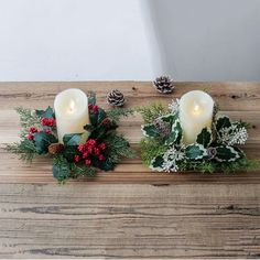 two white candles sitting on top of a wooden table covered in greenery and pine cones