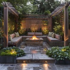 an outdoor fire pit surrounded by plants and seating with lights on the sides, in front of